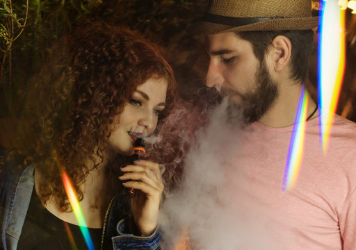 Loving couple on date. Guy and girlfriend smoke electronic cigarettes. He blows clouds of smoke. Red-haired girl. Man in hat and t-shirt. Lights of night city shine.