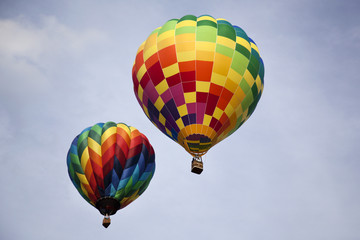 Two rainbow colored hot air balloons flying through the sky on a cloudy day.