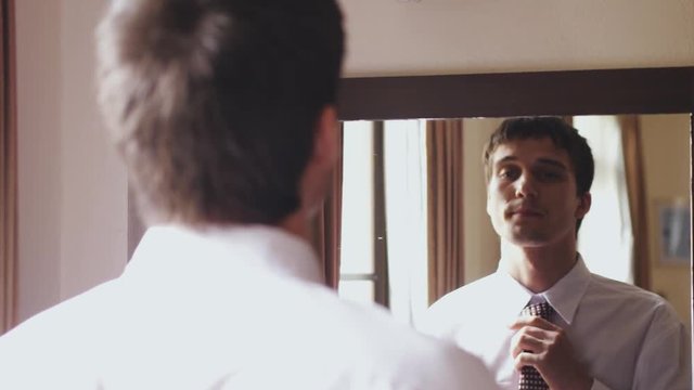 Young handsome man in white shirt stands by the mirror tying a tie. Changes focus from his back to his reflection in the mirror. 3840x2160, 4k