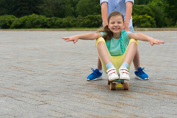 on a walk in the park, the boy pushes a girl in front of him, who sits on a skateboard