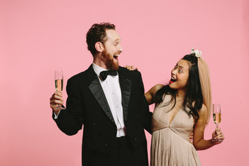 Bride and groom laughing and looking at each other while  holding champagne glasses, isolated on pink