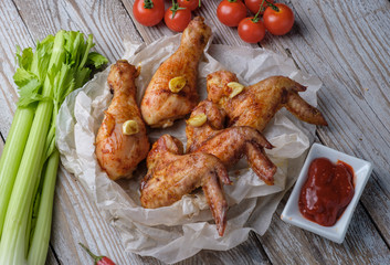 Baked chicken legs and wings lie on the table. Nearby are cherry tomatoes, celery, parsley and spices.
