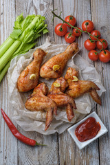 Baked chicken legs and wings lie on the table. Nearby are cherry tomatoes, celery, parsley and spices.
