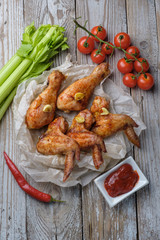 Baked chicken legs and wings lie on the table. Nearby are cherry tomatoes, celery, parsley and spices.