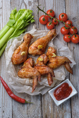Baked chicken legs and wings lie on the table. Nearby are cherry tomatoes, celery, parsley and spices.