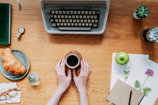 Stylish and modern wooden desk with typewriter, croissant,woman hands on cup of coffee notebooks, succulents and office accessories. Home office of romantic writer or poet. Flat lay concept.