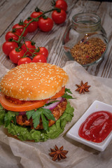 The hamburger lies on an old wooden table. On the table is a hamburger, tomato sauce, cherry tomatoes, a jar of spices.