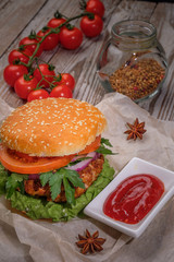 The hamburger lies on an old wooden table. On the table is a hamburger, tomato sauce, cherry tomatoes, a jar of spices.