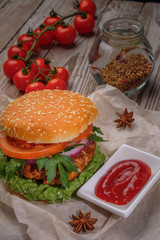 The hamburger lies on an old wooden table. On the table is a hamburger, tomato sauce, cherry tomatoes, a jar of spices.