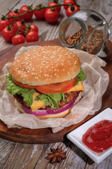 The hamburger lies on an old wooden table. On the table is a hamburger, tomato sauce, cherry tomatoes, a jar of spices.