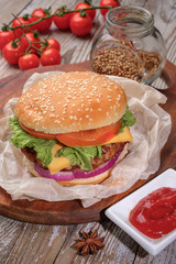 The hamburger lies on an old wooden table. On the table is a hamburger, tomato sauce, cherry tomatoes, a jar of spices.