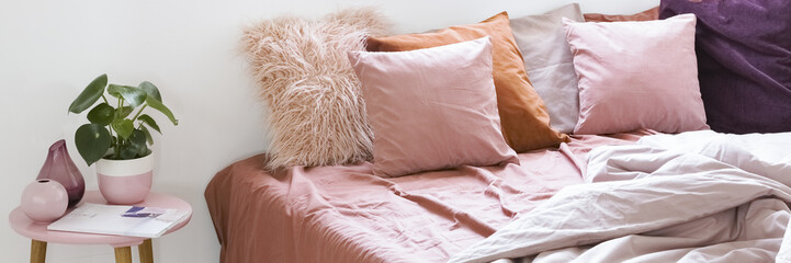 Real photo of a large bed with pink bedding and pillows standing next to a small table with a plant in bedroom interior - obrazy, fototapety, plakaty