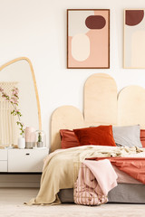 Posters above bed with red pillows in modern pastel bedroom interior with mirror. Real photo