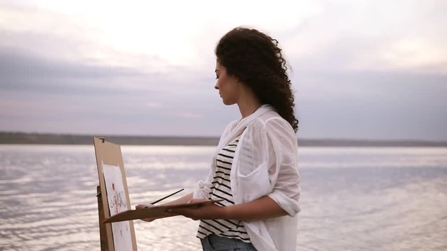 Outstanding view of a young curly brunette artist in working process, standing in the water with easel and drawing a picture with surrounding landscape. Outdoors