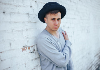 young handsome model man, young boy standing near white brick wall, wearing in black hat