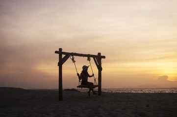 Fototapeta premium Young woman sitting on a swing at the beach with sunset sky