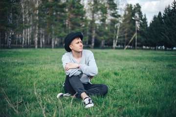 young man model in grey sweater rest sitting on grass in hat