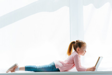 side view of cute little schoolgirl lying on windowsill and reading book