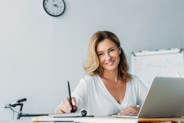 attractive businesswoman holding pen and looking at camera in office