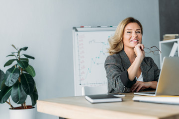 pensive smiling businesswoman holding glasses and looking away in office