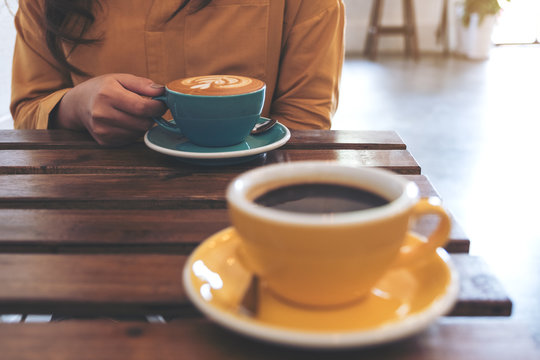 Closeup image of a woman holding a blue cup of hot coffee and another yellow cup on table in cafe
