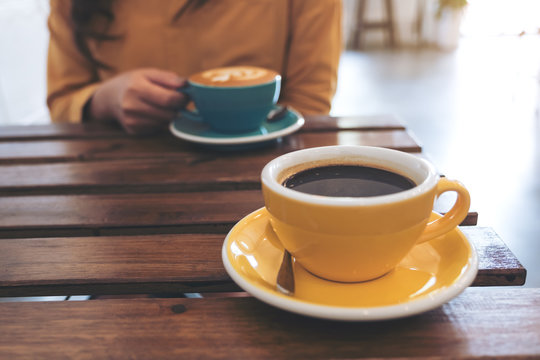 Closeup image of a woman holding a blue cup of hot coffee and another yellow cup on table in cafe