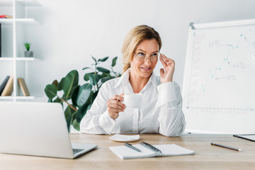 Obraz premium beautiful businesswoman holding cup of coffee and looking away in office