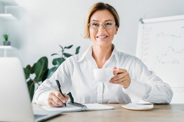 attractive businesswoman holding cup of coffee and writing to notebook in office