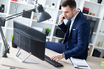 Fototapeta premium Młody mężczyzna siedzi w biurze na biurku, rozmawia przez telefon i pracuje z komputerem.