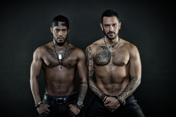 Fototapeta na wymiar Machos with muscular tattooed torsos look attractive, dark background. Guys sportsmen with sexy muscular torsos. Athletes on confident faces with nude muscular chests. Sexy body concept