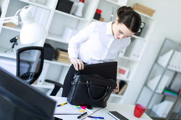 A young girl in the office is standing near the table and puts things in a bag.