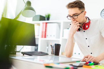 A young man in glasses stands near a computer desk. A young man draws a marker on a magnetic board. On the neck, the guy's headphones hang.