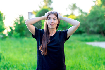 Beautiful brunette girl with long hair and black t-shirt. Summer in nature among green meadows. He holds head in his hands. She looks at the sky with a glance. Emotional dreams and fantasies.