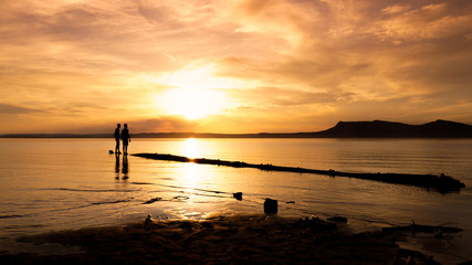 Black silhouette of people walking on a log in the water at a beautiful sunset