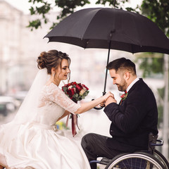 Bride and groom on the wheelchair sit kissing on the bench in the park