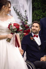 Bride and groom on the wheelchair hold their hands together standing before the altar