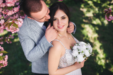 Beautiful, cheerful and lovely newlyweds embrace near the blooming pink cherry blossom, the groom straightens the bride's hair. Wedding portrait of a close-up of a smiling groom and a cute bride.