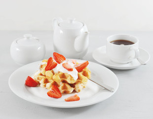 Belgian waffles with strawberries and cream. Next cup with tea, a teapot, a plate with fresh strawberries. Serving for breakfast. Light background. Close-up.