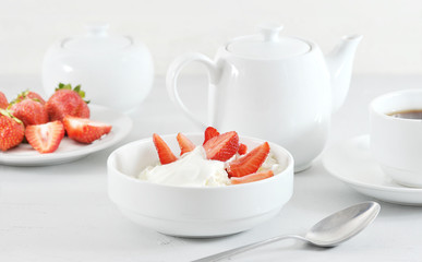 Cottage cheese with sour cream and fresh strawberries. Next cup with tea and a teapot. In the background is a plate of strawberries and a sugar bowl. Table setting for breakfast. Light background.