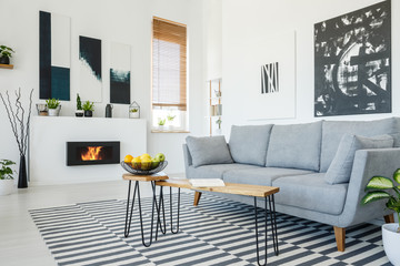 Real photo of a grey sofa standing in front of a wooden table in living room interior with posters on the walls, striped rug and bio fireplace