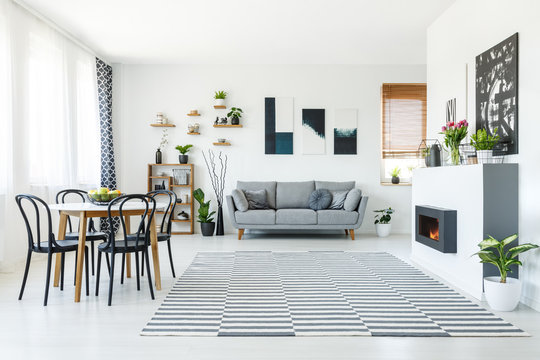 Real photo of a grey sofa standing in spacious living room with bio fireplace, table with chairs, wooden shelves, plants and paintings on the walls