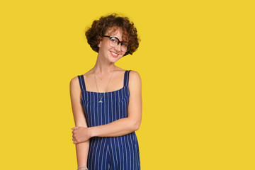People, beauty and lifestyle concept. Attractive sensual girl with curly brown hair and wide smile dressed in blue overall smiling broadly on yellow background. Joyful nice female