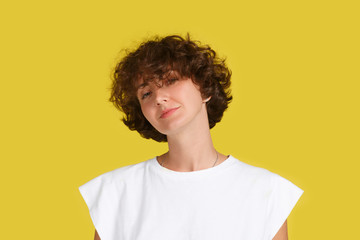 Portrait of pleased beautiful European female with curly brunette hair, dressed in casual white t shirt on yellow background. Attractive woman student has confident look. Facial expressions concept