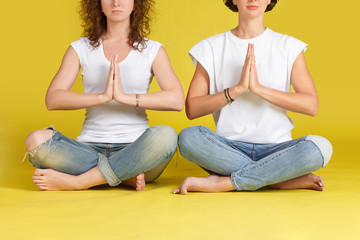 Fototapeta na wymiar Two Young curly woman practising yoga, holding hands in namaste and keeping eyes closed. Caucasian girls meditating indoors on floor at yellow background, having calm and peaceful facial expression