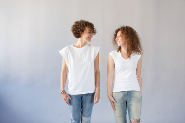 Studio shot of two European sisters with curly hair, wearing white t-shirts and jeans, smiling happily, looking at each other, posing at gray wall. People and friendship concept