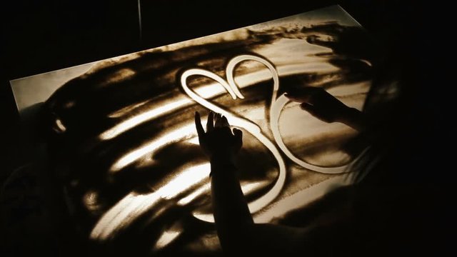Technology of drawing on the sand. The female artist is drawing two kissing swangs.