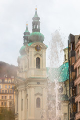 Obraz premium Hot spring geyser of mineral water in Karlovy Vary. Fountain with thermal water and church on background. Ancient european city in Czech Republic