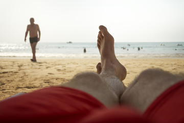 legs and feet of young male tourist lying on beach chair in front of water and blue sky with sun