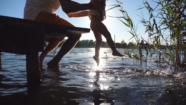 Young dad sitting on the edge of a wooden jetty at lake and holding his son in hands. Little child swinging feet in the water on a sunny day. Family spending time together outdoor. Side view Close up