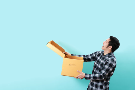 Side View Of Happy Customer Asian Man Smiling Opening And Holding Box Looking Up To Copy Space Above With Isolated Blue Background Feeling Excited And Satisfaction. Ordering Or Buying Concept Design.